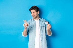 Jovem Fazendo Compras Online Com Aplicativo Movel Segurando Um Smartphone E Um Cartao De Credito Em Pe Sobre Um Fundo Azul Scaled